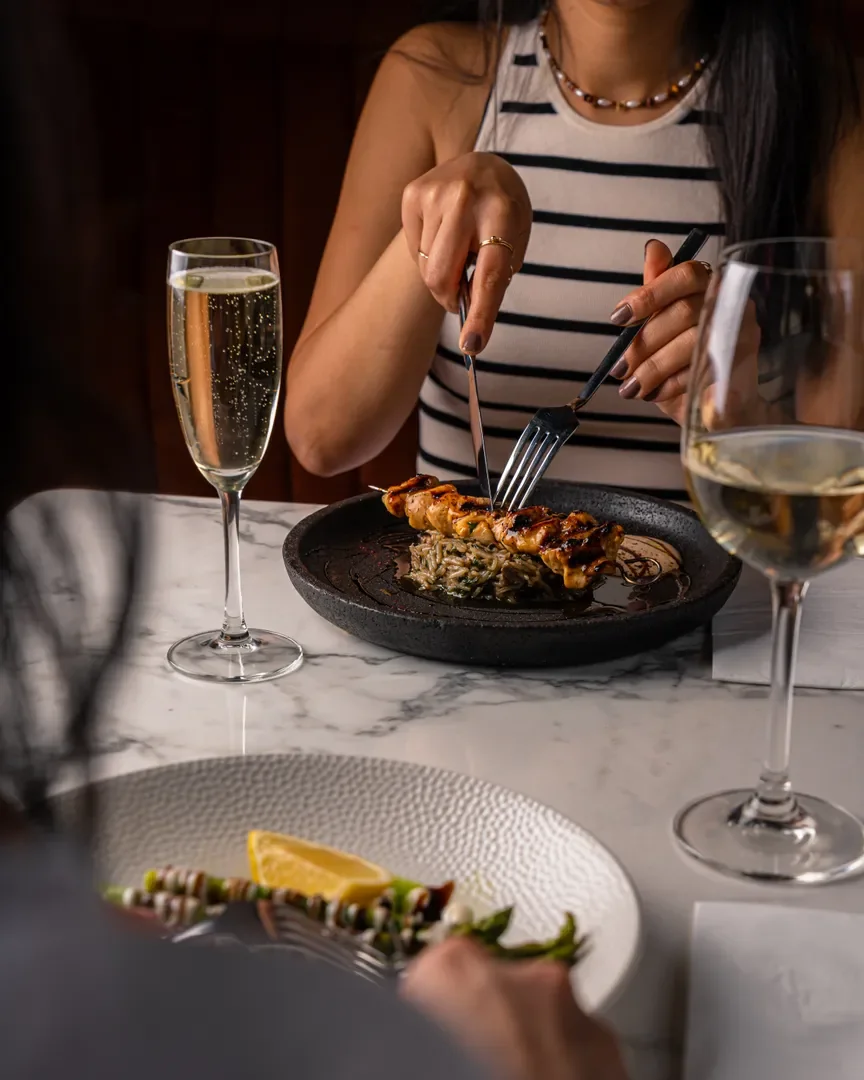 A glass of prosecco and a glass of white wine on a marble table, beside a grilled skewer